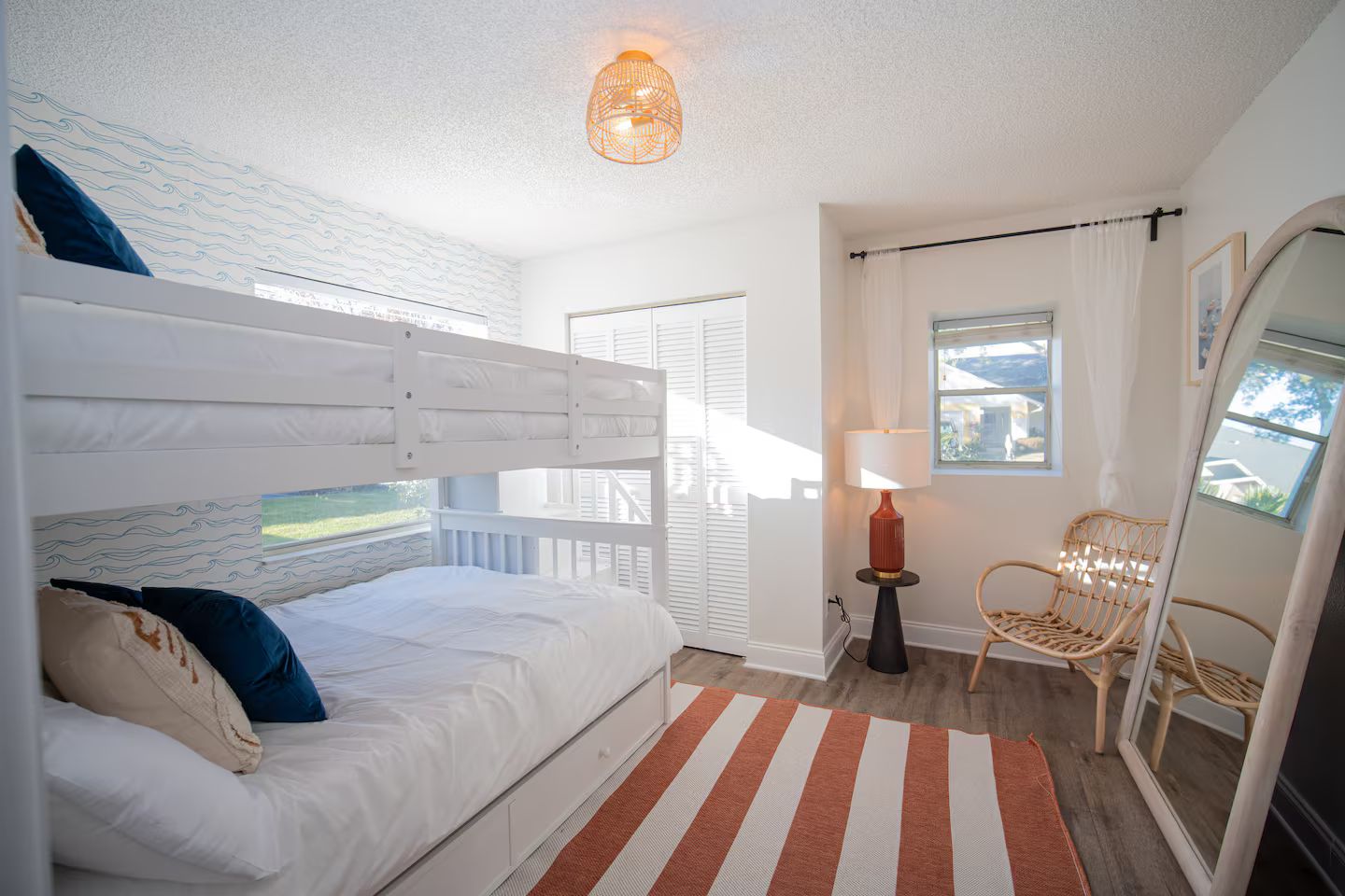 A bedroom with a bunk bed, a bedside lamp, a wicker chair, and a view through a window.