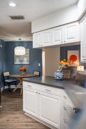 A modern kitchen with white cabinetry, a dining area with a table and chairs, and a vase with flowers on the counter. - Palm Harbor Heated Pool Resort - Theater Mini Golf (Palm Harbor)