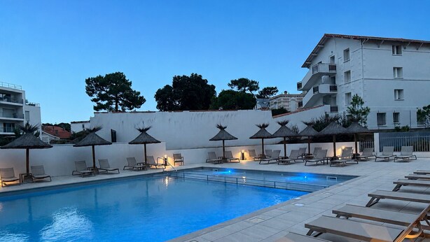 Piscina com espreguiçadeiras e guarda-sóis, cercada por um edifício com vários andares e varandas.