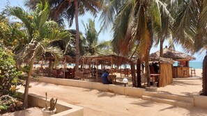 A beachside restaurant with outdoor seating, palm trees, and a thatched-roof hut. - Vrbo Property (M'bour)