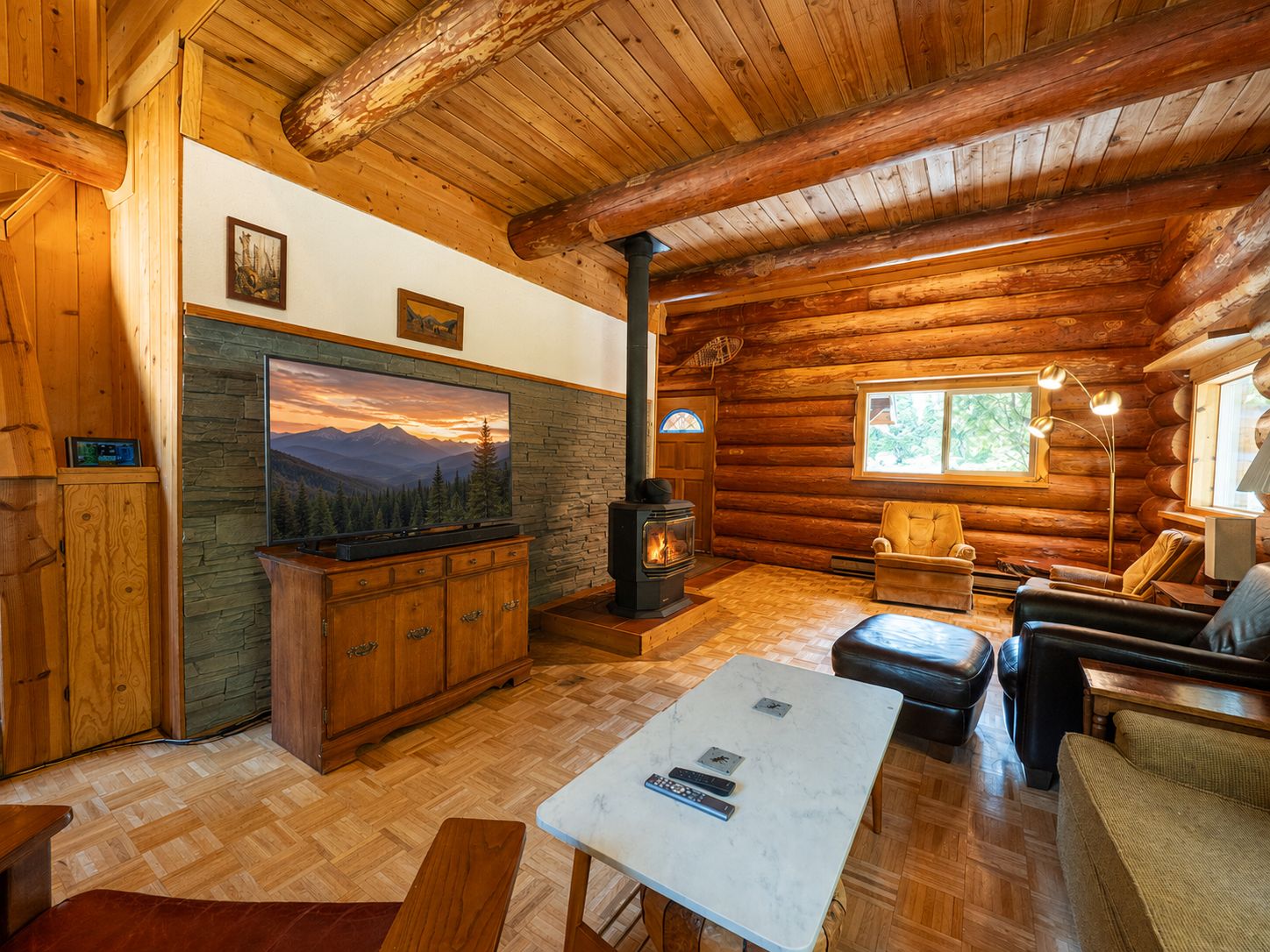 Interior de una cabaña de troncos con chimenea de piedra, muebles de madera y un cuadro grande de un paisaje montañoso.