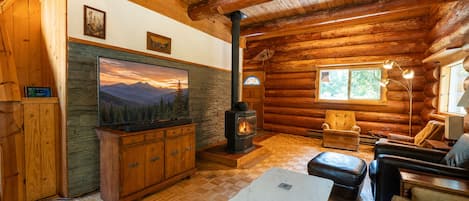 Interior de una cabaña de troncos con chimenea de piedra, muebles de madera y un cuadro grande de un paisaje montañoso.