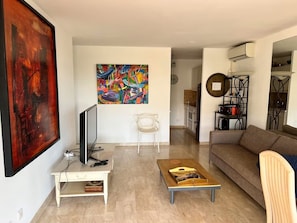 A living room with a television, a coffee table, a sofa, and a large abstract painting on the wall. - Apartment, Air-Conditioned, Ocean View, Large Pool, Locked Garage (Nice)