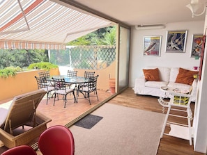 A room with a glass door leading to a patio, a white sofa, a small table, and a wall with framed artwork. - Apartment, Air-Conditioned, Ocean View, Large Pool, Locked Garage (Nice)