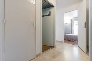 A hallway with a bed in the adjacent room, a door with a handle, and a wall with a cityscape mural. - Apartment, Air-Conditioned, Ocean View, Large Pool, Locked Garage (Nice)