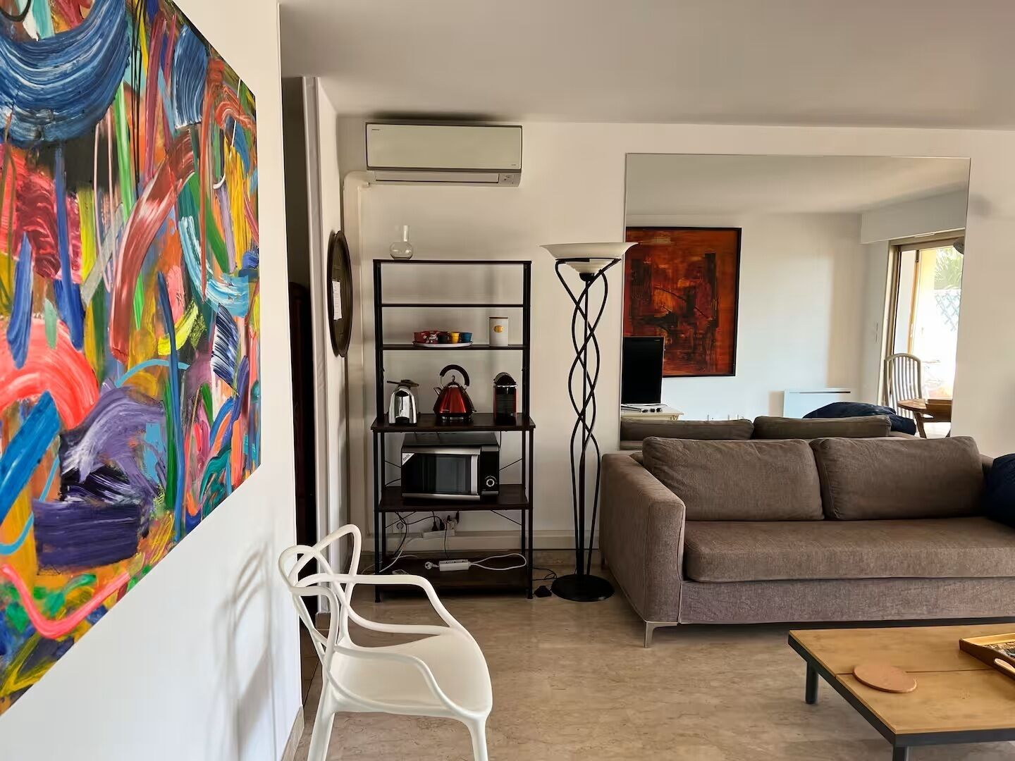 Una sala de estar con un sofá, una silla blanca, una lámpara de pie, un televisor y un cuadro abstracto colorido en la pared.