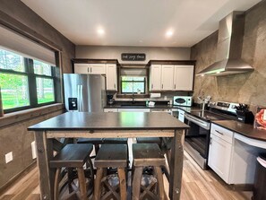 A modern kitchen with a central island, white cabinets, stainless steel appliances, and a view of greenery outside.