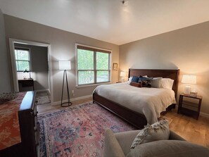 A bedroom with a large bed, two bedside tables with lamps, a floor lamp, a desk with a lamp, and a window with blinds. - Long Term | 1 BR/1.5BA | Communal Pool & Hot Tub (Whitefish)