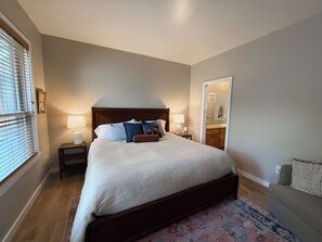 Quarto com cama grande, mesas de cabeceira, cômoda, espelho e janela com persianas.