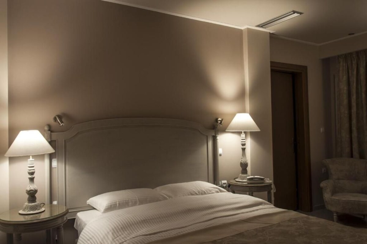 Quarto de hotel com uma cama grande, duas mesas de cabeceira com luminárias, uma cadeira e uma porta.