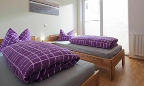 A room with two beds, each with a purple checkered bedspread and matching pillows. There is a nightstand with a lamp on one side and a window with blinds on the other. - Holiday Apartment in Norderney With Wi-fi (Norderney)