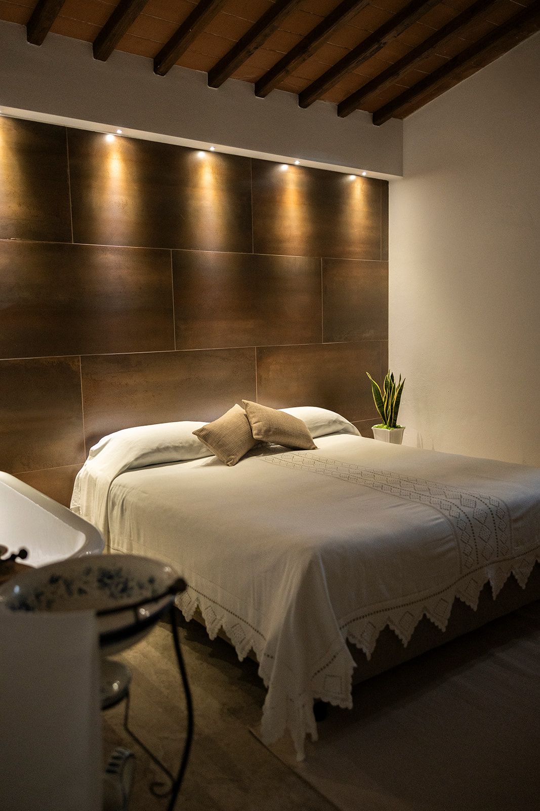 A bedroom with a modern headboard, a bed with white bedding, a small potted plant, and a bedside table with a lamp.