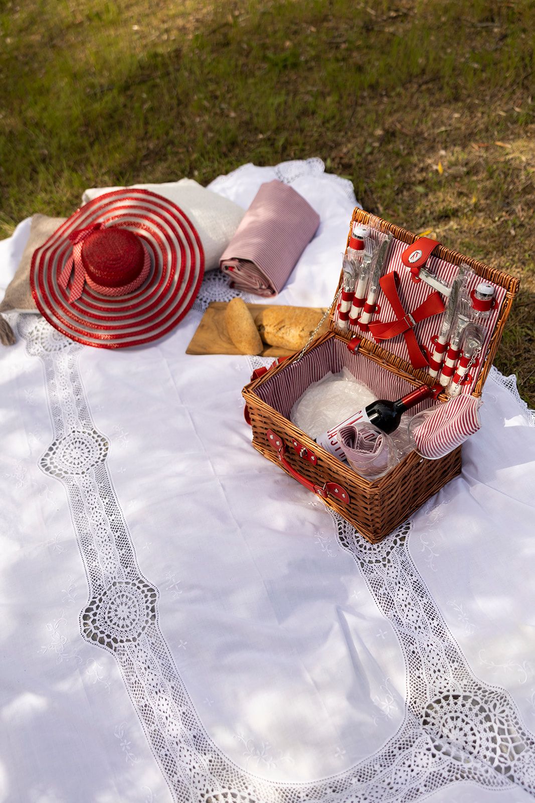 A picnic setup with a basket containing bottles, a rolled-up cloth, and a hat on a white lace tablecloth.