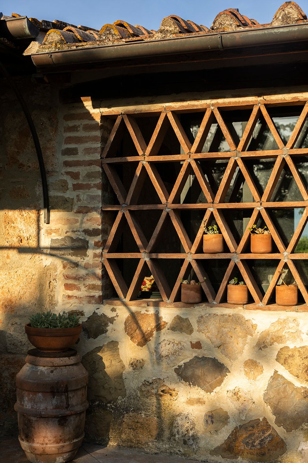 A rustic stone wall with a metal lattice window, potted plants, and a large metal barrel.