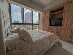 A bedroom with a large bed, a TV mounted on a wooden cabinet, and a view of buildings through the window. - Avmx Living 5A 1 bed (Guadalajara)
