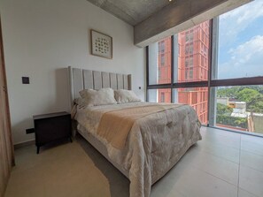 A bedroom with a bed, a nightstand, and a large window with a view of buildings.