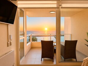 Uma varanda com duas cadeiras de vime, uma TV na parede e vista para o pôr do sol sobre o oceano.