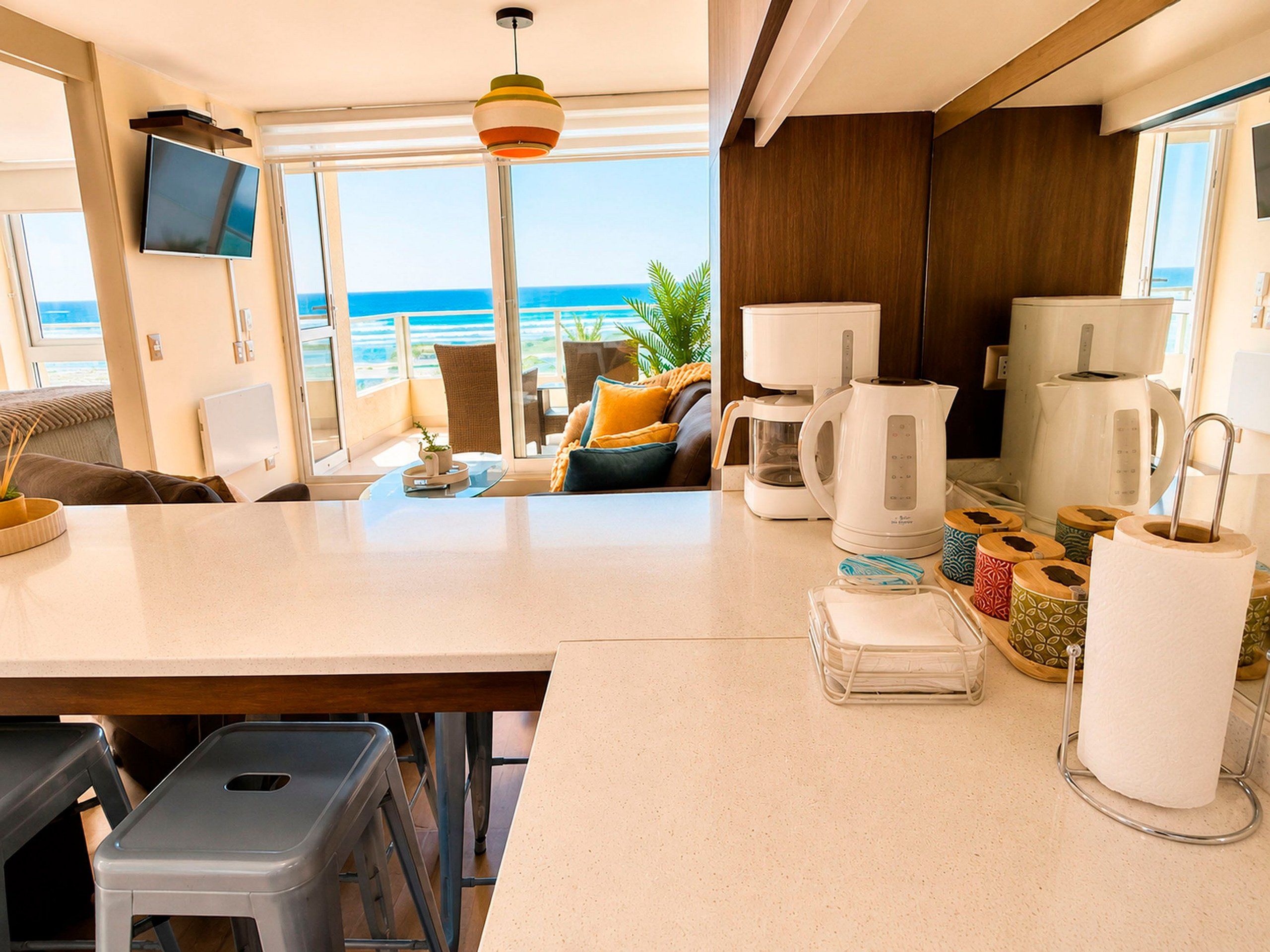 A modern kitchen with a view of the ocean, equipped with a coffee maker, kettle, and a TV mounted on the wall.