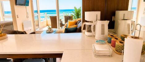 A modern kitchen with a view of the ocean, equipped with a coffee maker, kettle, and a TV mounted on the wall.