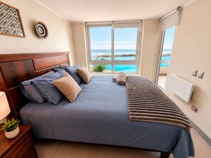 A bedroom with a bed, a nightstand, a lamp, a potted plant, a window with a view of the sea, and a radiator. - Laguna del Mar ☆ The Sea from Your Terrace (La Serena)