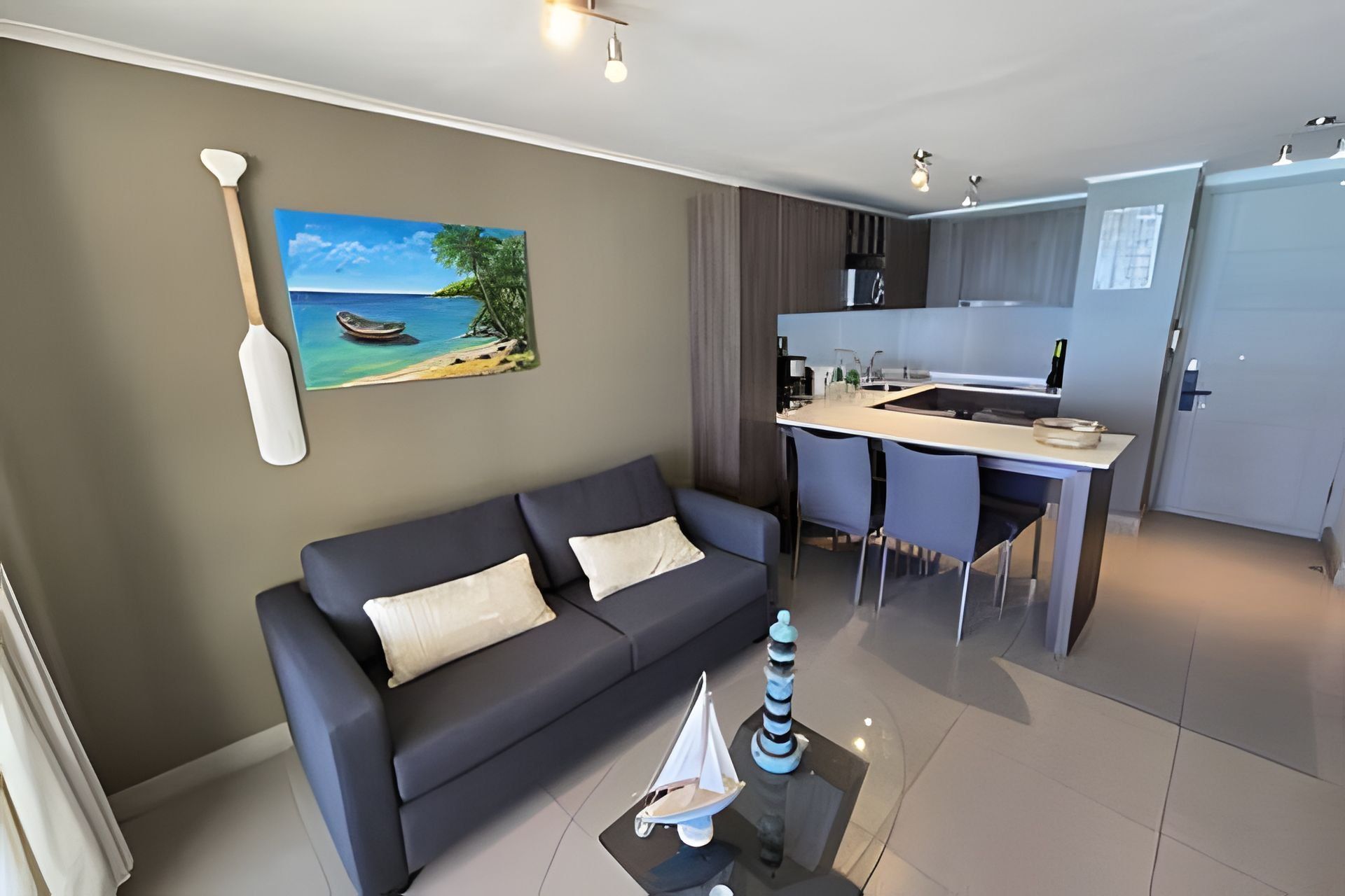 Un salon moderne comprenant un canapé, un tableau représentant une plage, une petite maquette de voilier et un coin cuisine avec une table à manger.