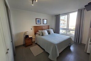 Ein Hotelzimmer mit Bett, Nachttisch, Lampe, einem Fenster mit Vorhängen und Blick auf Gebäude.