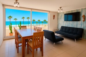 Ein Wohnzimmer mit großem Fenster zum Strand, einem Essbereich mit Holzküchenmöbeln und einem schwarzen Sofa.