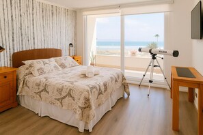 Ein Schlafzimmer mit einem Bett, einem Nachttisch, einem Schreibtisch mit einem Teleskop und Blick auf den Strand.