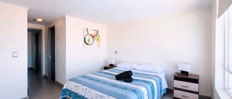 A hotel room with a bed, a bedside table, a lamp, a painting on the wall, and a window.
