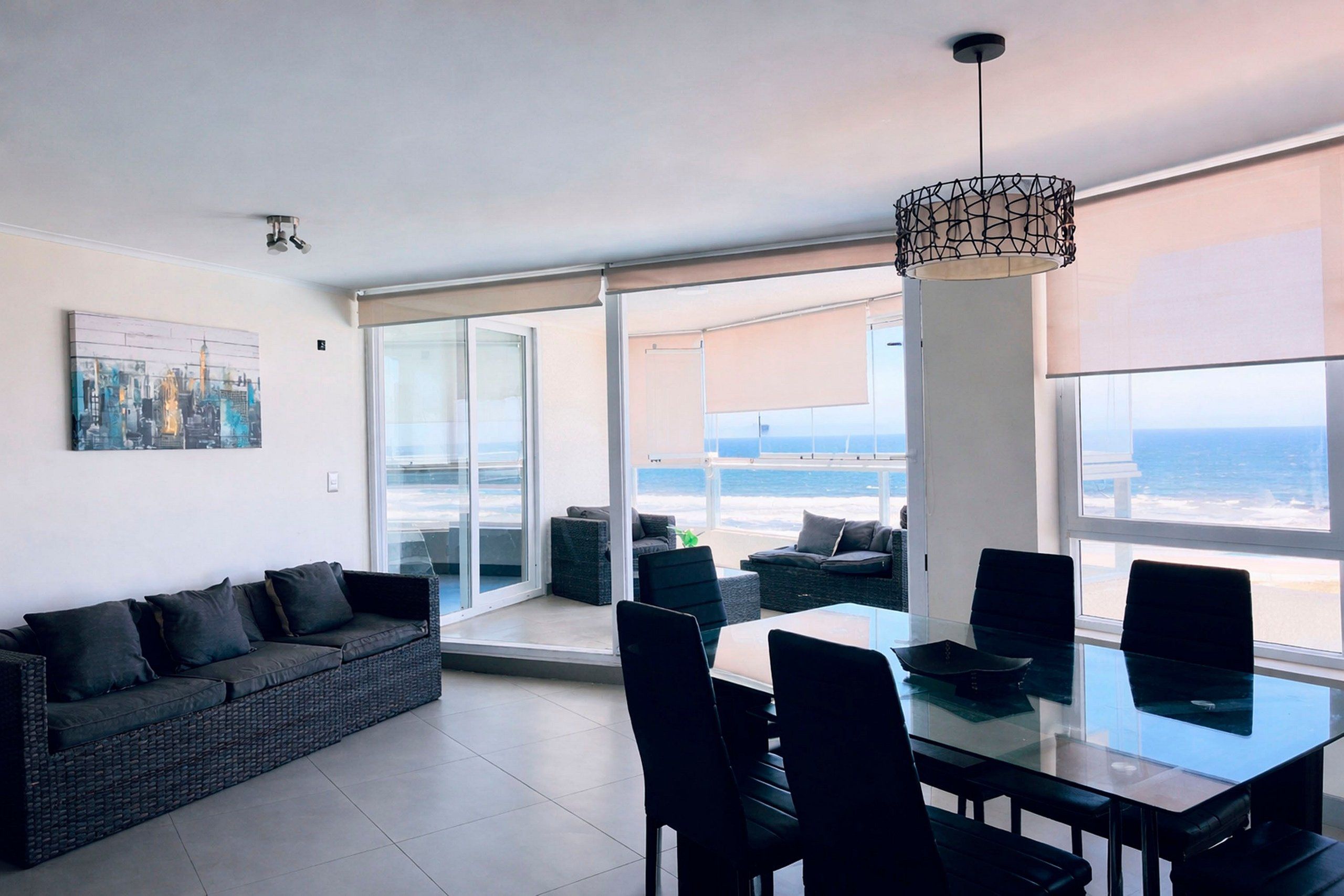 Een moderne woonkamer met een glazen tafel, zwarte stoelen, een grijze bank en een groot raam met uitzicht op zee.