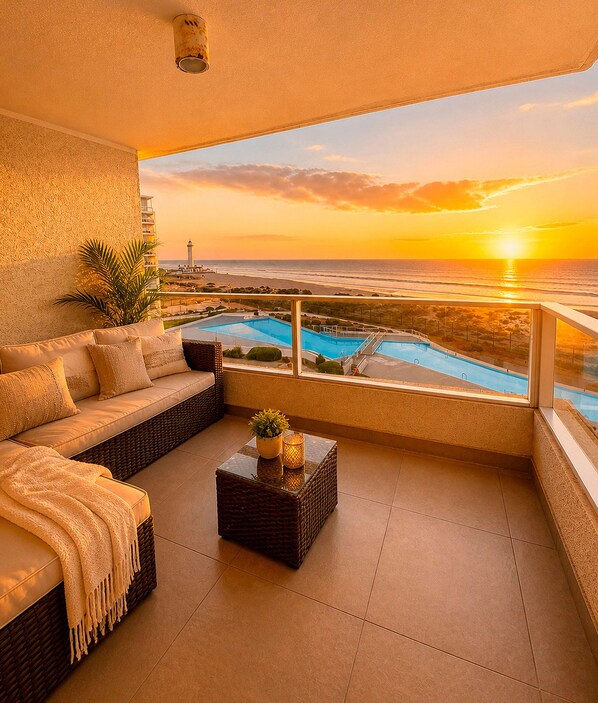 A balcony with a sofa, a small table, and a view of a swimming pool and sunset.
