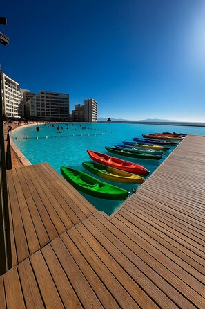 Une terrasse en bois, ornée de kayaks aux couleurs vives alignés, offre une vue sur une mer bleue et limpide, avec des bâtiments qui se dessinent à l’horizon.