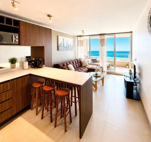 Une cuisine moderne dotée d’un îlot central et de tabourets de bar, un coin repas avec une table en verre, et un salon avec un canapé et de grandes fenêtres donnant sur la plage.