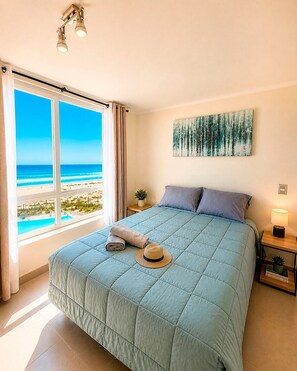 Une chambre avec une grande fenêtre donnant sur une plage, un lit recouvert d’une couette bleue, un chapeau et des serviettes enroulées posées sur le lit.
