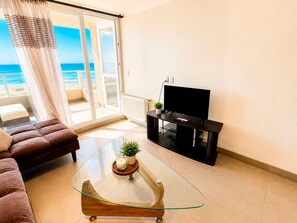 Ett vardagsrum med en soffa, ett soffbord i glas, en tv på ett tv-bord och utsikt över stranden genom en skjutdörr.