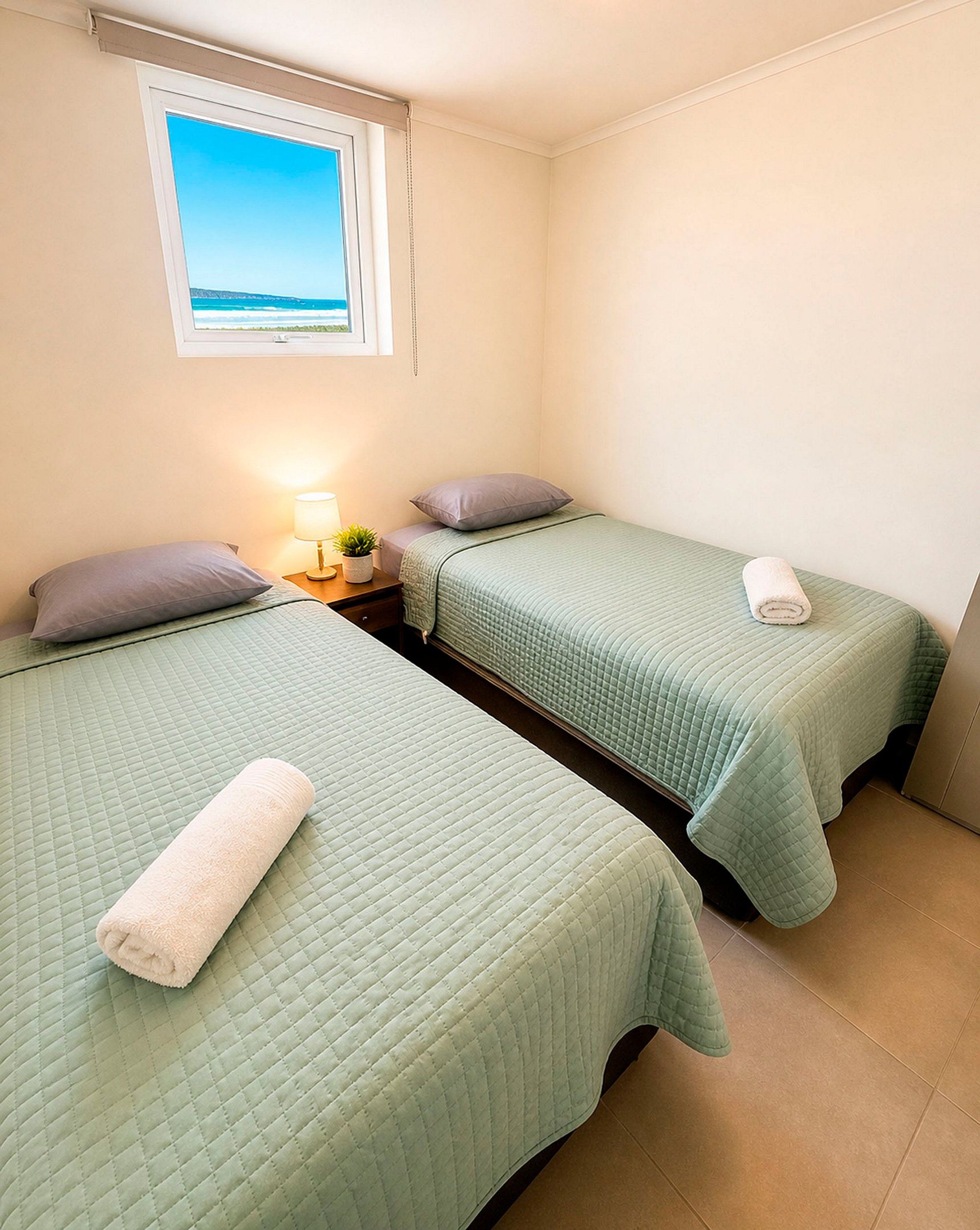 Une chambre double avec deux lits, une table de chevet avec une lampe et une fenêtre donnant sur la mer.