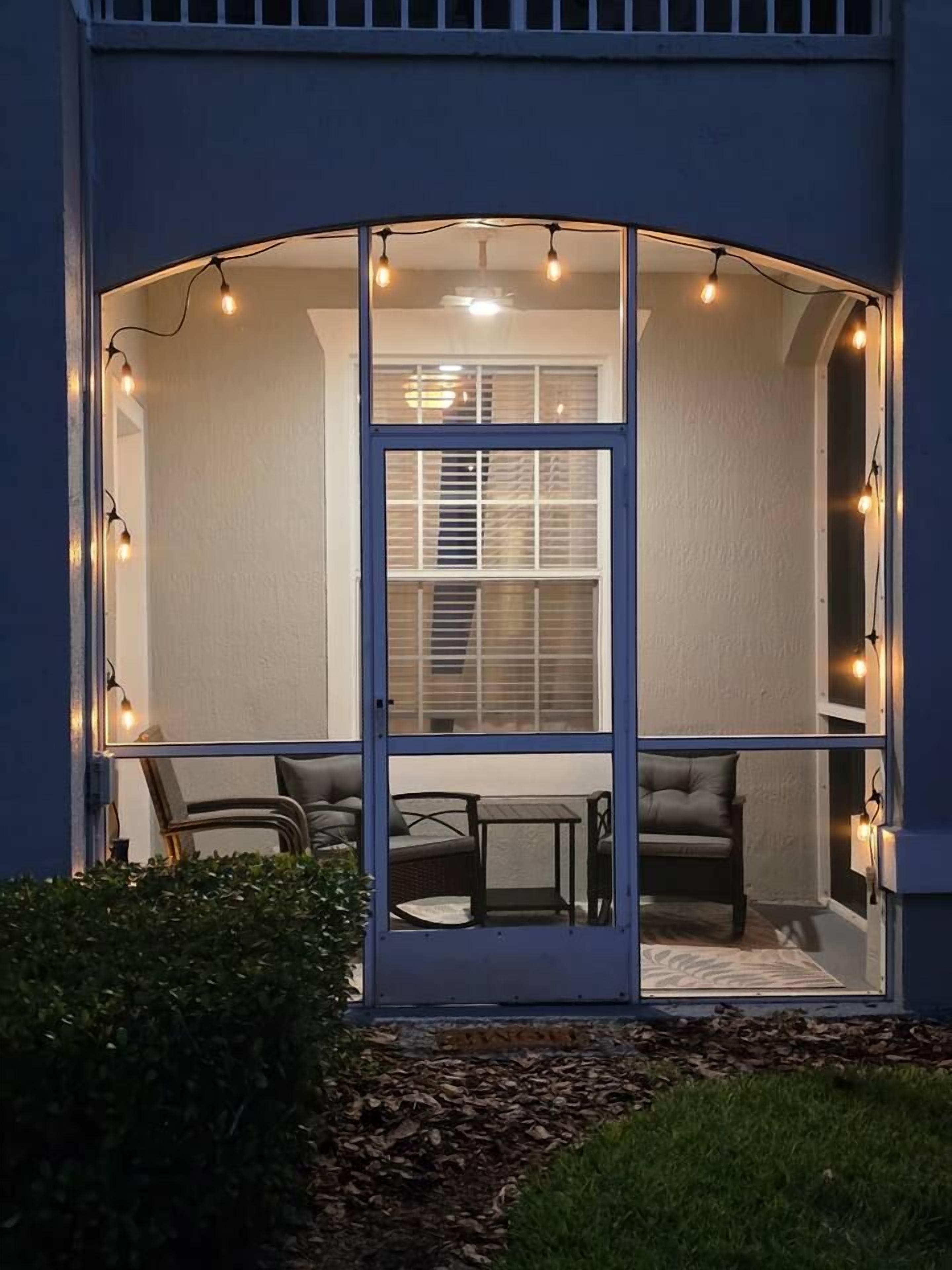 A glass-enclosed patio with string lights, a table, and two chairs.