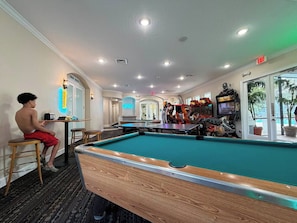 A pool table in a room with a pool, a bar area with arcade games, and a person sitting at a table. - Disney Escape at Windsor Palms (Kissimmee)