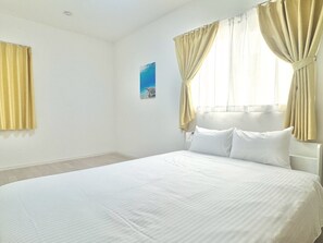 A bedroom with a bed, curtains, a painting, and a bedside table. - Vrbo Property (うるま市)
