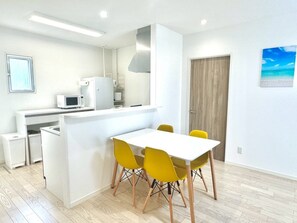 A modern kitchen with white appliances, a dining area with yellow chairs, and a wooden door. - Vrbo Property (うるま市)