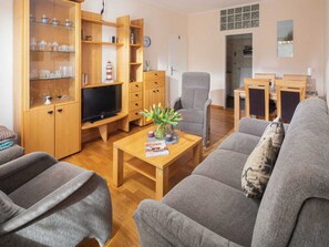 A living room with a sofa, armchairs, a coffee table, a TV, wooden shelving with glass doors, and a small plant. - Holiday Apartment in Norderney With Wi-fi (Norderney)