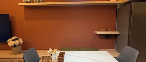 Ένας χώρος τραπεζαρίας με ένα λευκό τραπέζι, δύο καρέκλες, έναν πάγκο, ένα φωτιστικό οροφής και ένα ράφι με διακοσμητικά αντικείμενα.