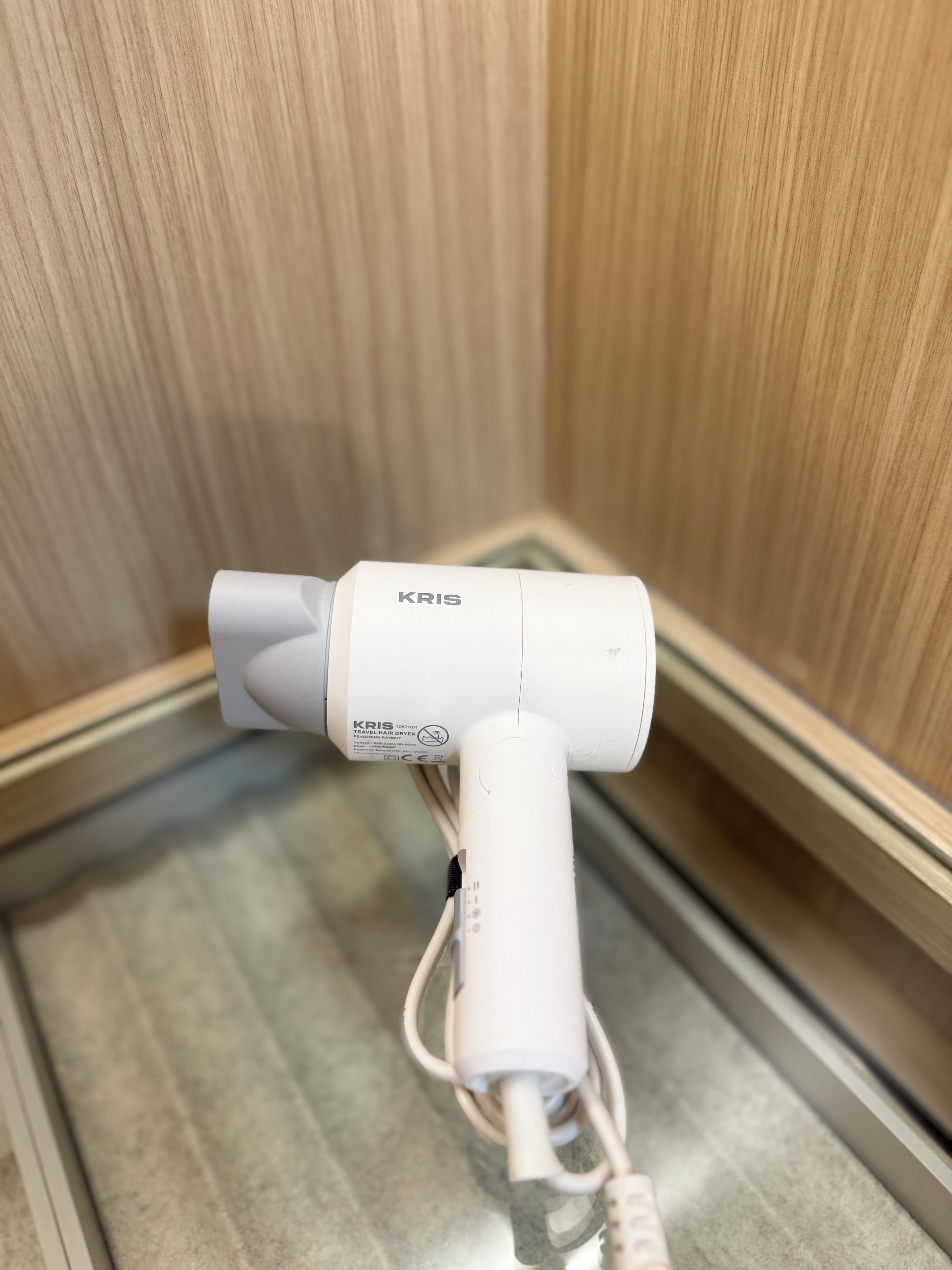 A white travel hair dryer with a cord, placed on a metallic surface against a wooden wall.