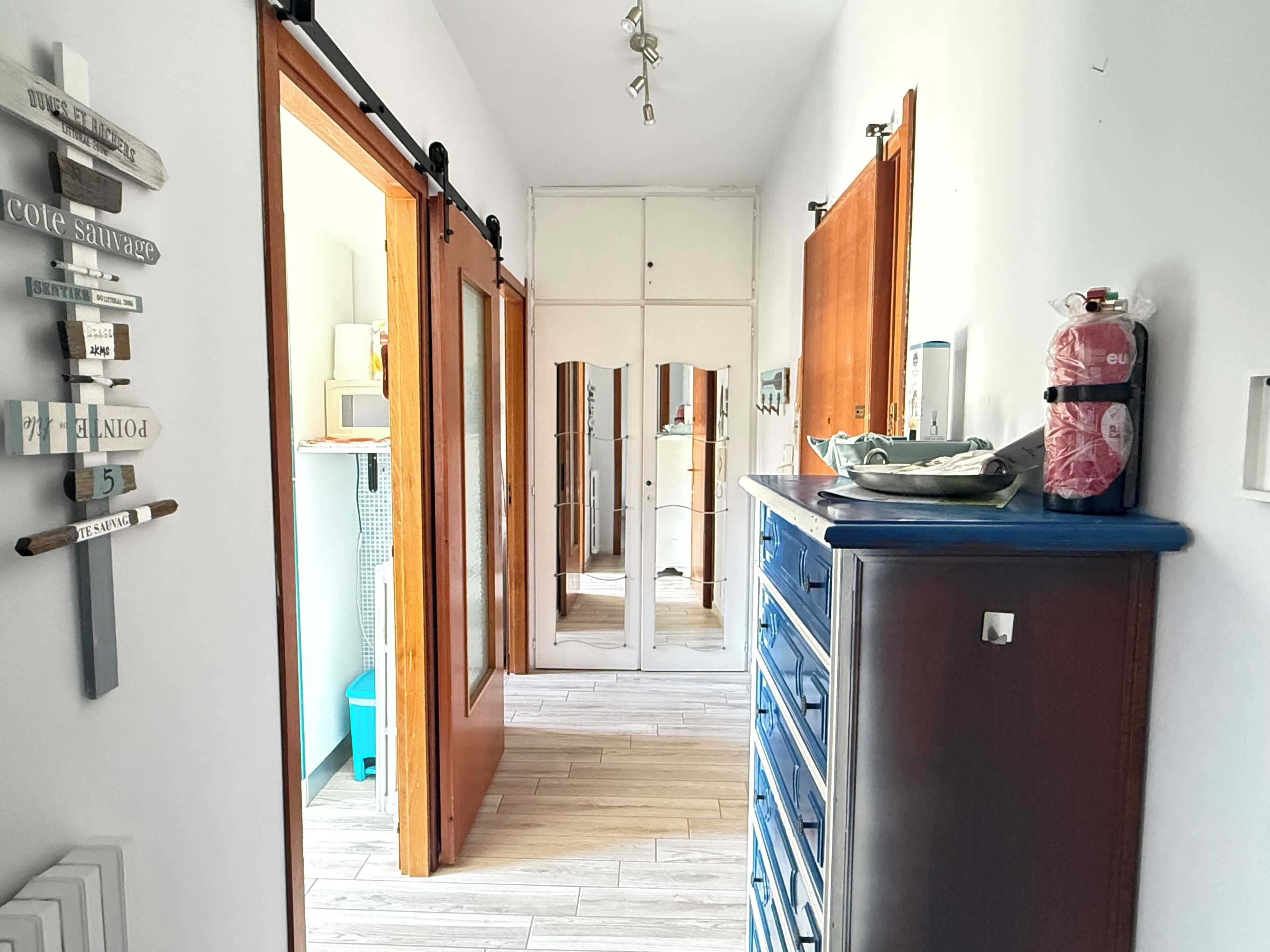 A hallway with a wooden floor, a kitchen area with cabinets, and a refrigerator.