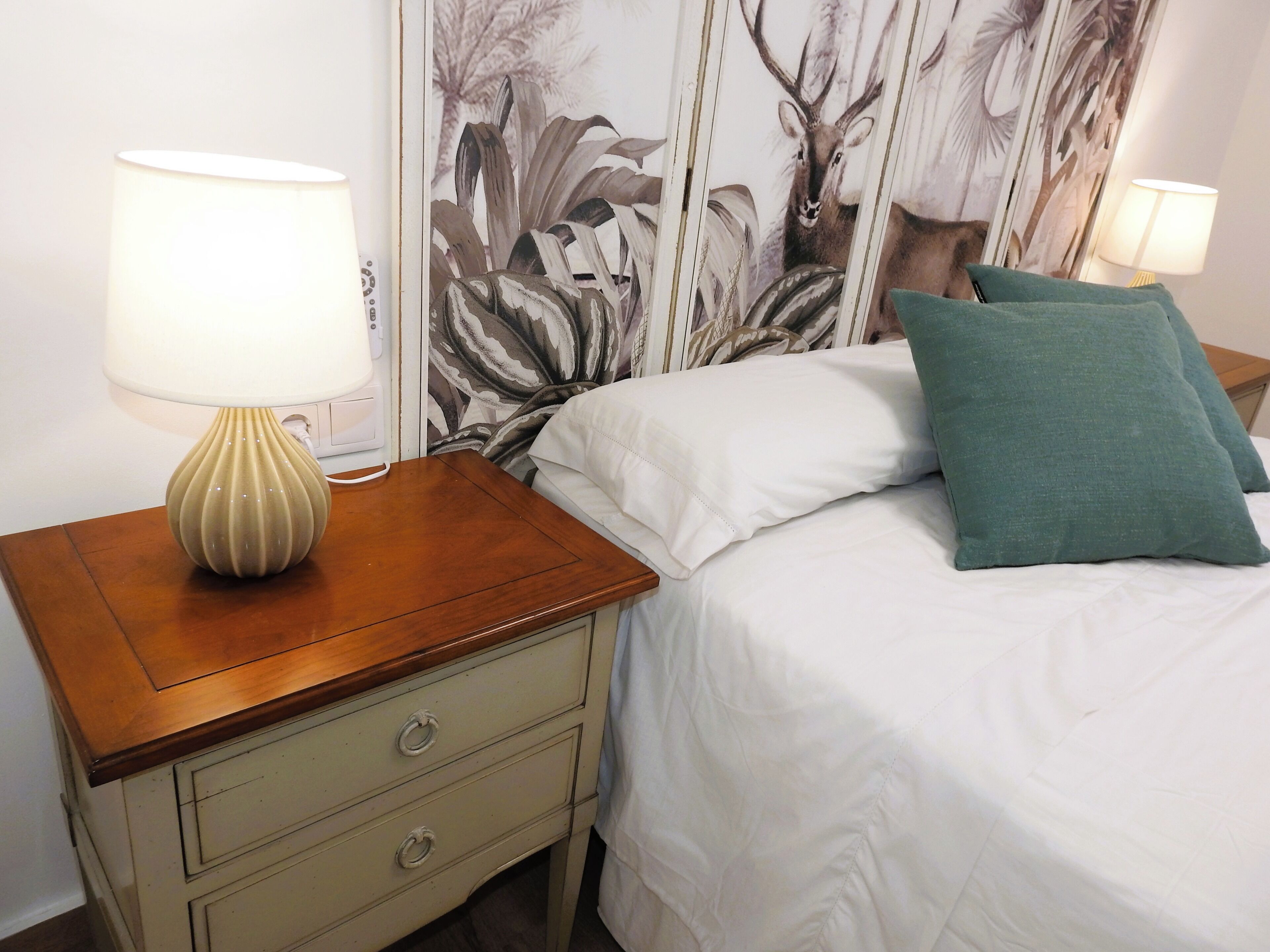 Een net opgemaakt bed met witte lakens en een turquoise kussen, een bedlamp op een houten nachtkastje, en een wand met een natuurprint.