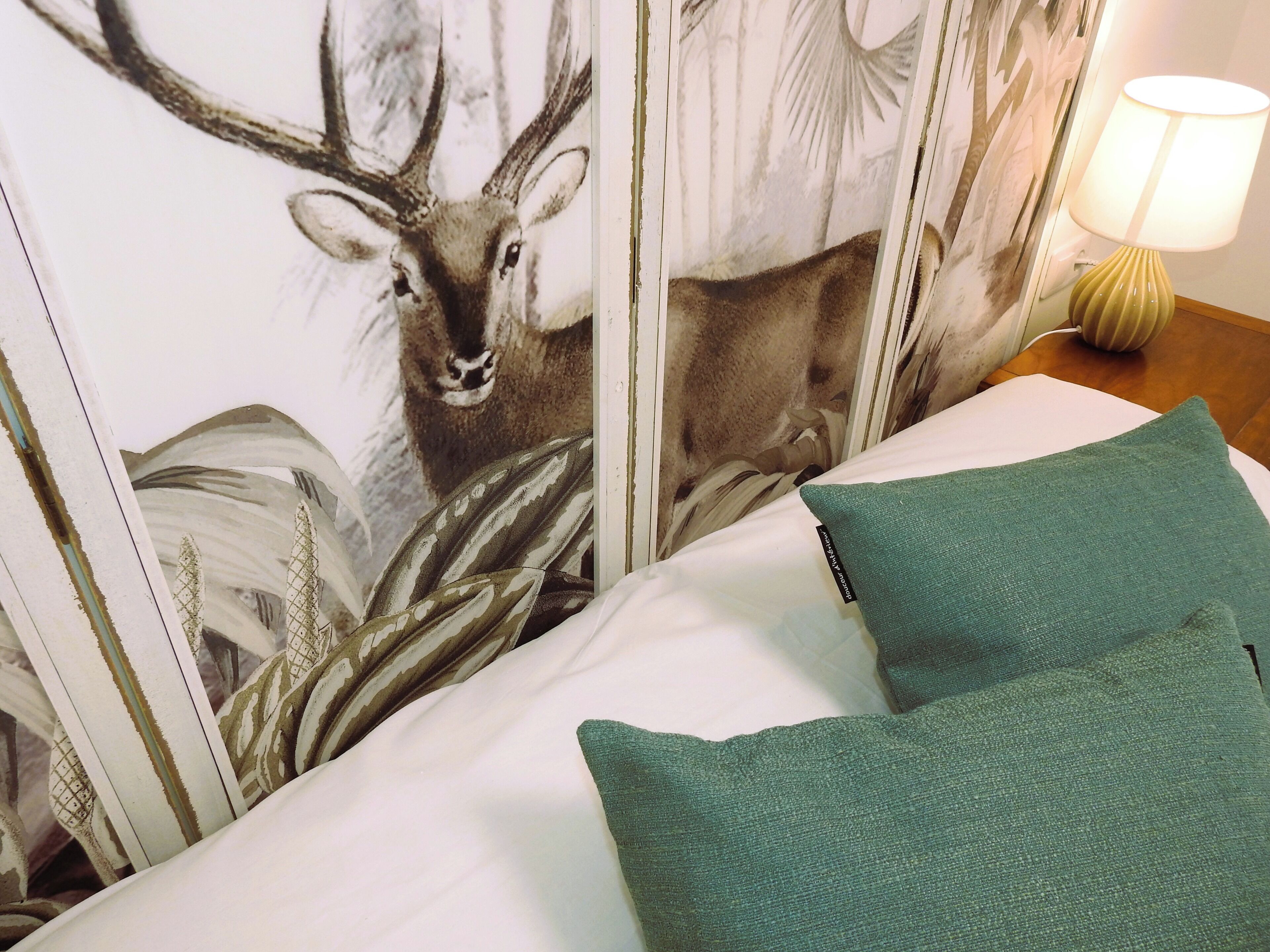 Quarto de hotel com uma cama, almofadas verdes e um mural de um veado na parede.