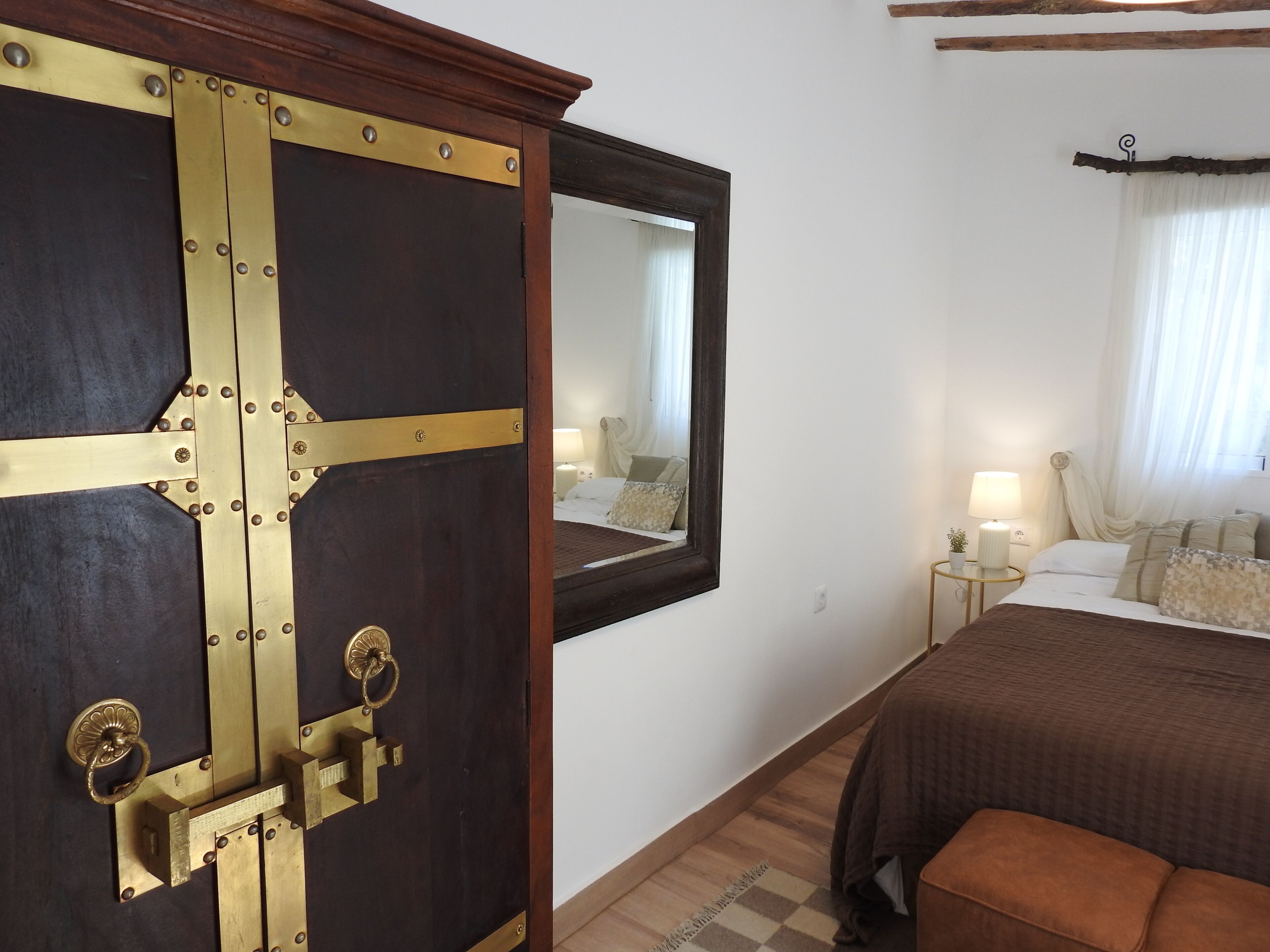 A hotel room with a large wooden door, a bed with a brown bedspread, a nightstand with a lamp, and a window with curtains.