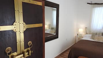 A hotel room with a large wooden door, a bed with a brown bedspread, a nightstand with a lamp, and a window with curtains.