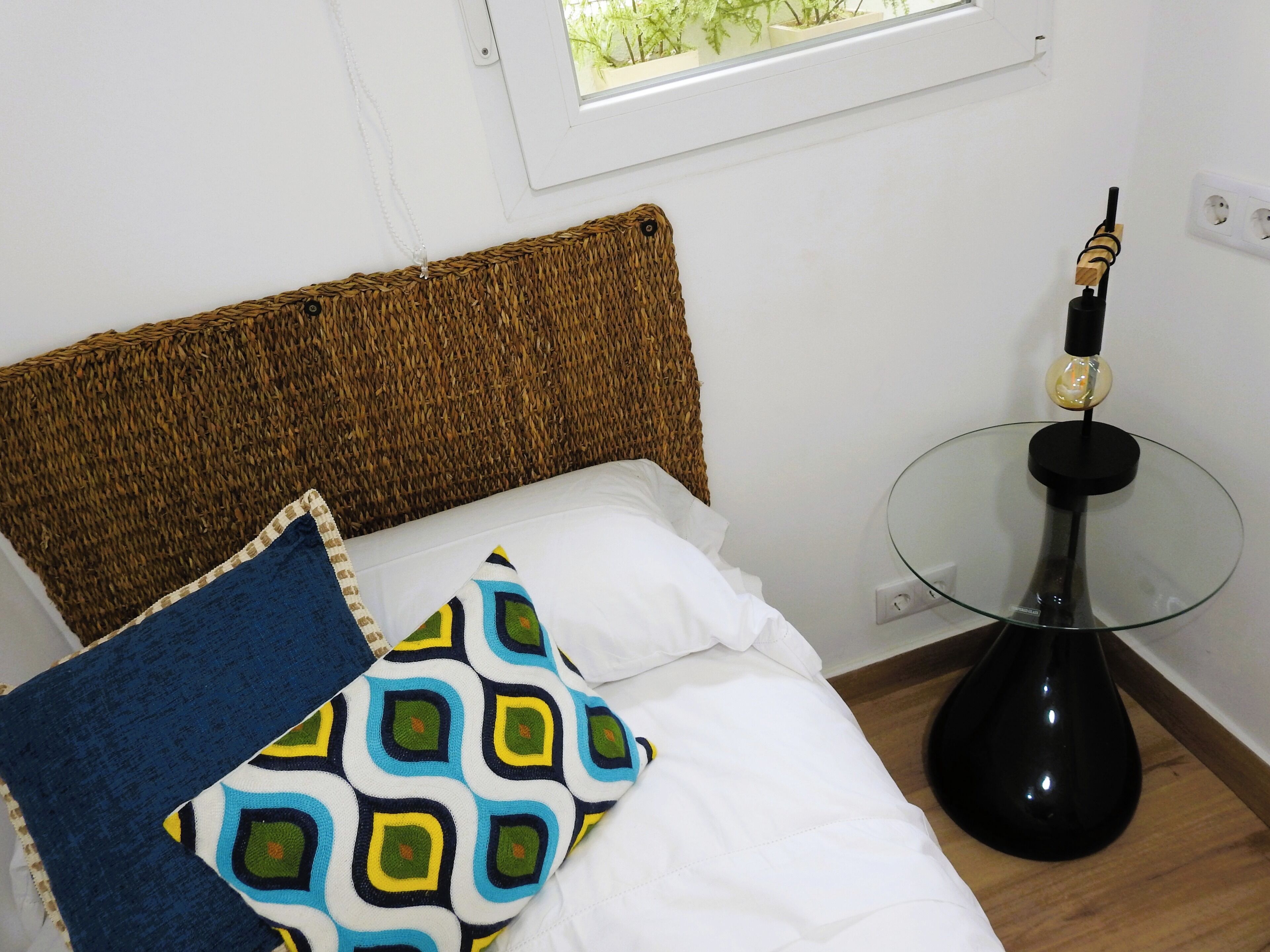 Un lit avec une tête de lit en osier, un coussin à motifs et une table en verre agrémentée d’un luminaire.