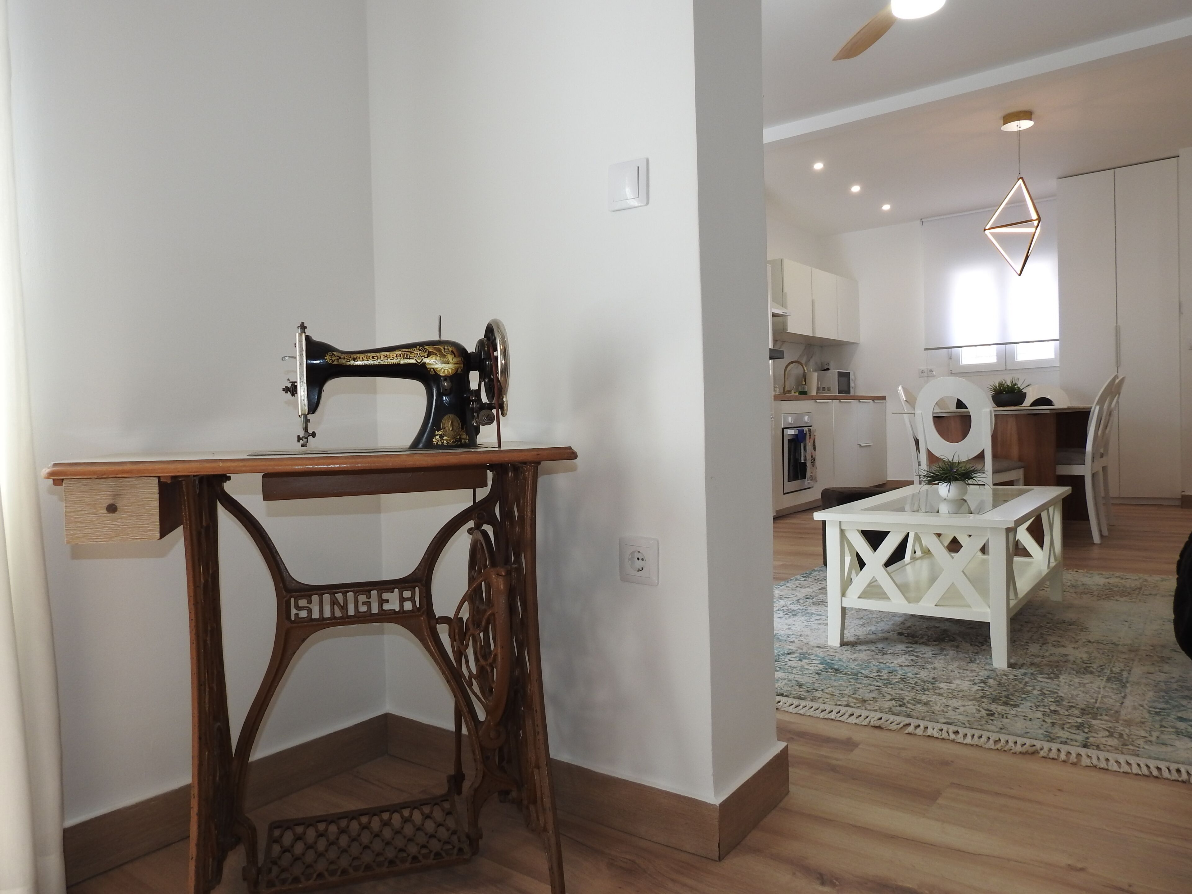 A sewing machine on a wooden table in a room with a kitchen in the background.